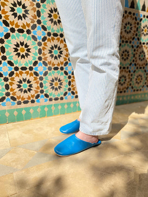 Babouches en cuir bleu faites main portées dans un riad marocain, mosaïque en arrière-plan