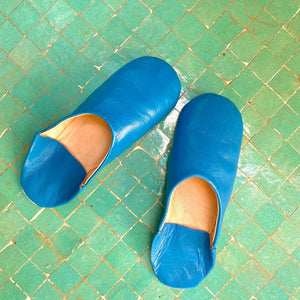Paire de babouches marocaines en cuir bleu posées sur un sol en zellige vert